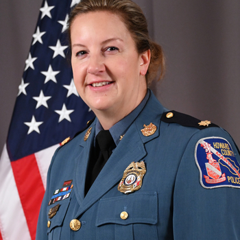 Major Cory Zirk, in uniform, standing in front of an American flag with grey background.